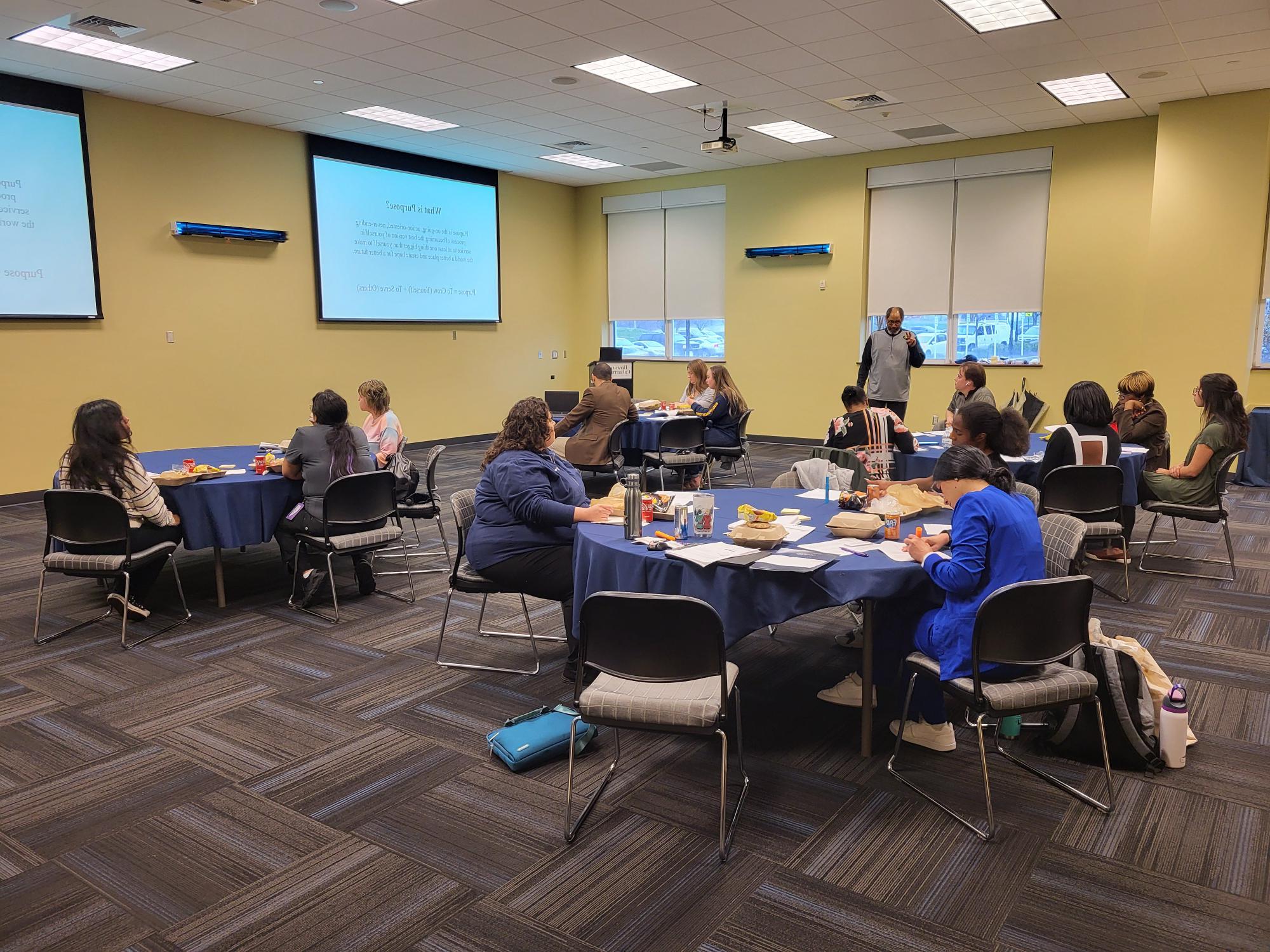 A conference room with attendees seated at round tables, a speaker presenting, and slides displayed on screens.